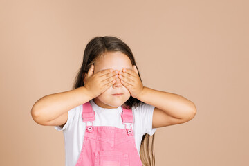  kid closing eyes with hands not see others with closed mouth not talk with others wearing bright ,pink jumpsuit and white t-shirt on beige background.