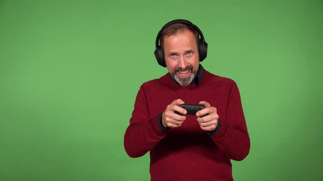 A middle-aged handsome Caucasian man plays a game on the camera, holds a controller and wears headphones - green screen background