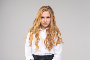 Portrait of young girl in white shirt with frowning face looks at camera on gray background. Caucasian blonde with curly hair is not happy with something.