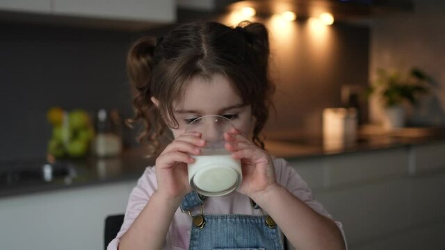 Little child drinking milk from a glass cup in her kitchen. Milk source of calcium for children.