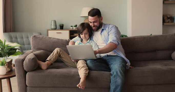 Caring father spend time at home with happy child son preschooler rest on couch teach kid to use app browse information online on modern laptop. Young dad little kid boy play video game on pc computer