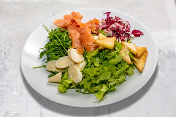 Mixed salad with smoked salmon, lettuce, rocket, red chicory, heart of palm and croutons. In white dish in top view