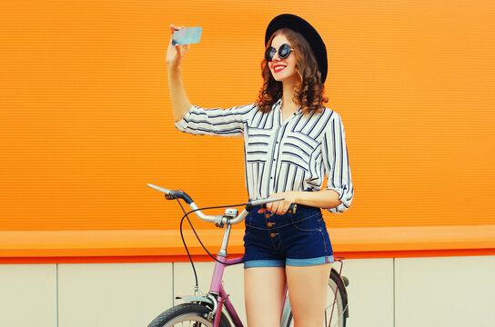 Beautiful happy smiling young woman taking selfie by smartphone with bicycle on orange background