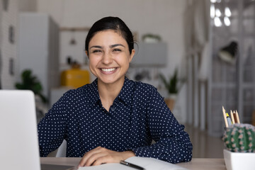 Happy mixed race Indian student girl looking at camera, smiling, working at laptop, studying from...