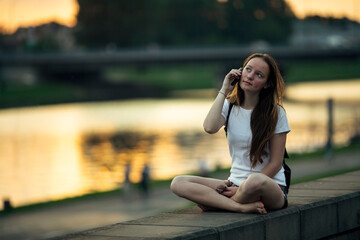 A girl talking on the phone while sitting on the city's waterfront at sunset.