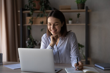 Fototapeta premium Happy pretty student girl watching online training, writing notes, smiling at screen. Millennial woman studying in business school from home, attending virtual class, remote lesson, learning webinar