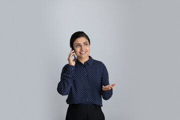 Happy young Indian businesswoman talking on mobile phone, looking away, standing by white...