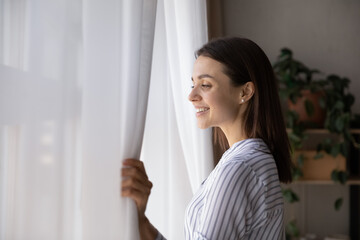 Happy young woman looking out of window at home, pulling back white curtains, smiling. Female renter, homeowner breathing fresh air in cozy apartment, enjoying leisure time, weekend, relaxing
