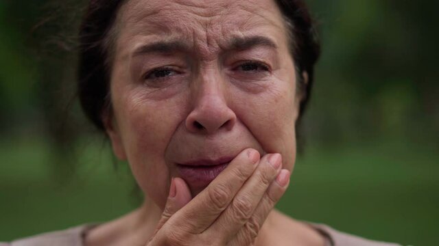 Headshot portrait crying desperate frustrated Caucasian senior woman closing face with hands. Close-up grieving hopeless female retiree looking at camera posing outdoors in park
