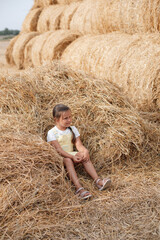 Big pile of hay harvest on field with young girl resting in it looking away with serious face holding hay in hand wearing sundress. Having fun away from city on field full of golden hay.