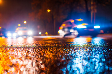 Night city in the rain.  Headlights of approaching cars. Reflections of light on a wet road. Falling drops of rain. Colorful colors. Focus on the asphalt. Close up view from the asphalt level.