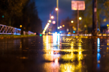 Fototapeta na wymiar Night city in the rain. The light from the lanterns reflects on the wet pedestrian sidewalk. Falling drops of rain. Colorful colors. Focus on the asphalt. Close up view from the asphalt level.