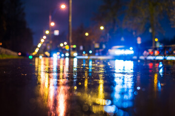 Night city in the rain. The light from the lanterns reflects on the wet pedestrian sidewalk. Falling drops of rain. Colorful colors. Focus on the asphalt. Close up view from the asphalt level.