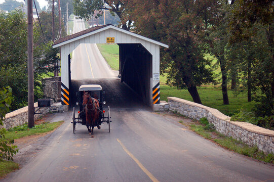 An Amish horse and buggy emerge from a covered bridge as it travels the landscape and country lanes of Lancaster County, Pennsylvania