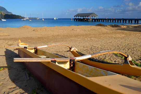 An outrigger canoe and kayak rests on a tranquil beach within site of a fishing pier on Hanalei Bay, Kauai
