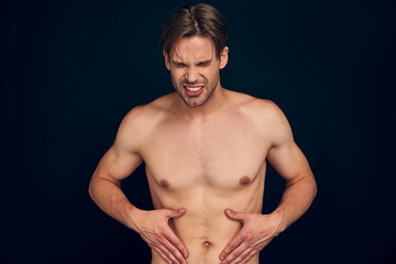 Fototapeta na wymiar Handsome young bearded man isolated. Cropped image of shirtless muscular man is standing on dark blue background. Man holding his stomach with big scars. Experiencing stomach pain