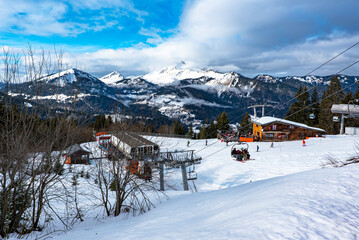 Skiers going up the mountains by the chair lift with a wooden lodge and high snow covered Alps mountains on the background. France 2022. - obrazy, fototapety, plakaty