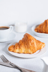 Croissant in bright white environment, with cup of coffee and milk and other croissant in blurry background. Simple and elegant breakfast setting and scene.