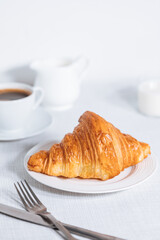 Croissant in bright white environment, with cup of coffee and milk and other croissant in blurry background. Simple and elegant breakfast setting and scene.