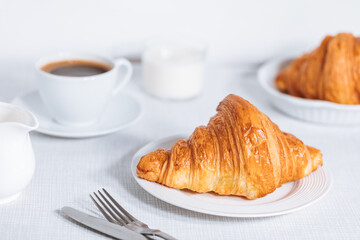 Croissant in bright white environment, with cup of coffee and milk and other croissant in blurry background. Simple and elegant breakfast setting and scene.