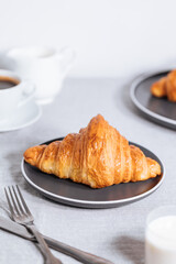 Croissant in bright white environment, with cup of coffee and milk and other croissant in blurry background. Simple and elegant breakfast setting and scene.