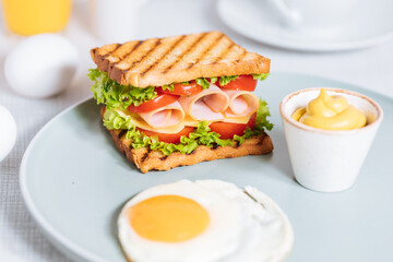 Toast sandwich with gouda cheese and turkey ham filled with tomato and lettuce on a bright background with ingredients in blurry background. Served with coffee and orange juice and eggs