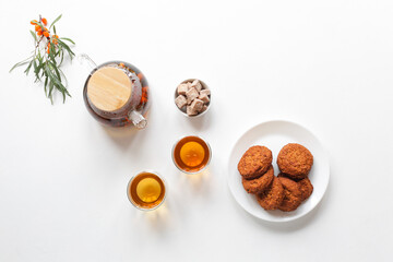 Healthy tea, adaptogenic drink, herbal tea with sea buckthorn berries. On a white table, a glass teapot and cups, cereal cookies with dried fruits on a wooden board. Top view, copy space.
