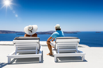 A elegant couple on sunbeds enjoys the view of the blue sea and sky of the Caldera at Santorini...