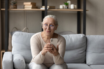 Happy beautiful mature old retired woman in eyeglasses with smartphone in hands looking in distance, distracted from communicating in social network, thinking of received message with good news.