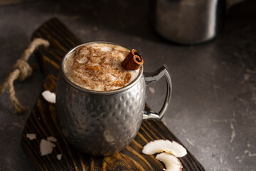Making coffee or cocoa  (latte, cappuccino) with foam in metal rustic mug witch cinnamon stick and powder on wooden board with coconut. Winter spiced hot drink, copy space. Close up dark photo.