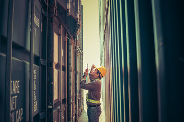 Asian man worker, standing between containers, using walkie-talkie radio and looking up at the containers, to worker and large transport in the export industry concept.