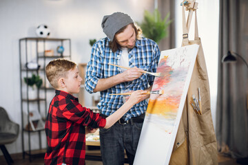 Handsome man and little boy in checkered shirts drawing abstract pattern on canvas. Happy father and son using brush and colorful paint during art process.