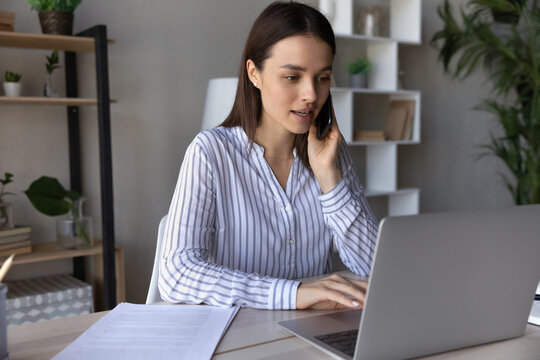 Serious young female employee speaking on telephone and using app laptop at office workplace. Multitasking manager talking on mobile phone, making call to client, advisor giving consultation