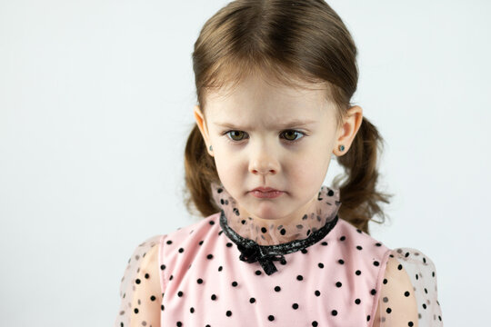 A little girl with two ponytails in a dress with polka dots on a white background looks down angrily. Studio photo