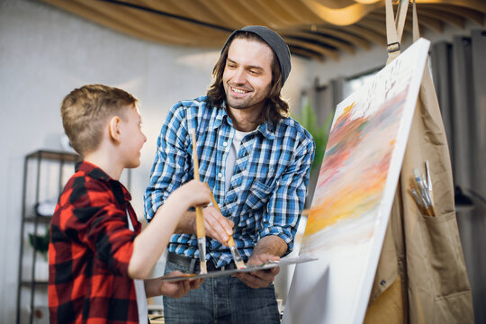 Handsome man and cute boy painting with brush on easel at home. Loving father and son looking on each other with sincere smile on face and enjoying time spending together at home.