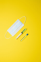 mask and syringe filled with a medicine, items lined up diagonally on a yellow background