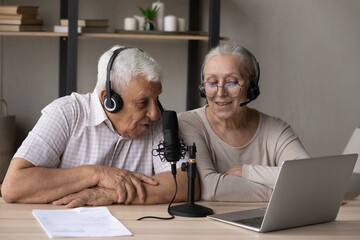 Happy middle aged old 70s family couple in headphones speaking in professional stand microphone,...