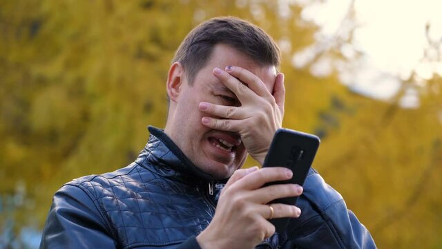 Sad millennial guy looks at phone screen in surprise and stands outside at autumn. Disgusted and overwhelmed young man stares at screen of smart phone, needs to pay debts immediately