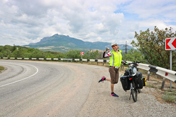 Obraz premium Happy woman bike traveler stands near a loaded bike on the highway among the mountains.