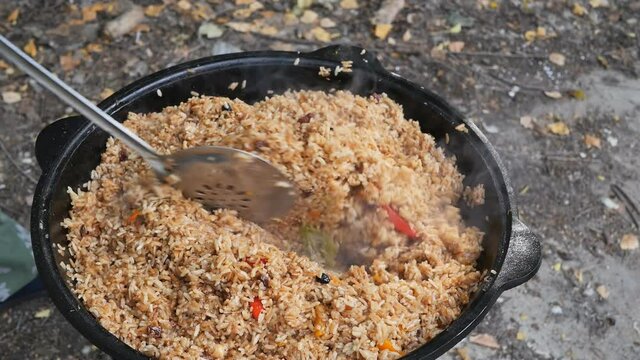 a man cook is stirring a ready-made hot pilaf in a cauldron with a slotted spoon and walks around. Full readiness of the Uzbek pilaf in the open air. Slow motion