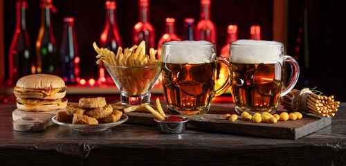 Glass of beer on bar counter. Jugs, mugs, pints of brew beverage, ale, cider on wooden table in pub, bar. Backlit dark showcase with craft beer bottles in brewery. French fries fried potatoes snack