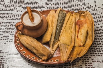 Sweet pineapple and blue tamales and red chili tamales, accompanied by white masa atole. Typical Mexican food. On a clay plate and accompanied by corn and cinnamon.