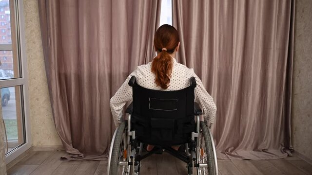 Caucasian woman in a wheelchair drives up to the window and opens the curtains. 