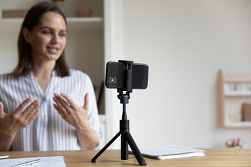 Positive trendy blogger woman speaking at smartphone webcam on tripod, recording post for vlog....