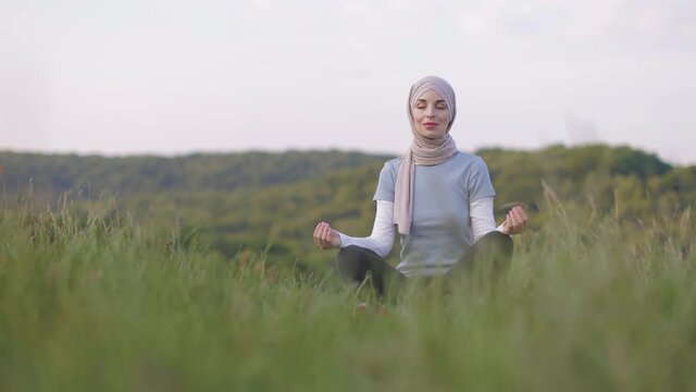 Peaceful young lady in hijab meditating with closed eyes in the park. Pretty Muslim woman sitting on couch in yoga pose, finding inner balance