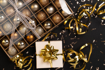 Holiday gift box or present with ribbon, golden confetti and gold baubles on black background. Magic New Year greeting card. Christmas Decoration. Mock up. Flat lay. 