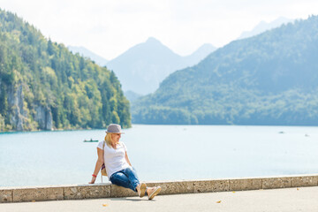 Fototapeta na wymiar Beautiful tourist lady wearing hat and backpack enjoying stunning view on Bavarian mountains near Neuschwanstein castle in Germany. Traveling concept. Nature view