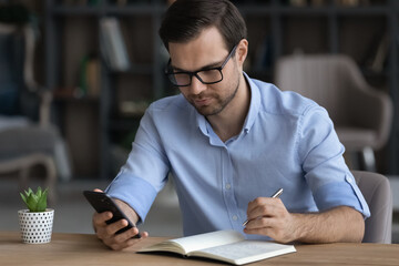 Focused confident businessman in glasses holding smartphone, taking notes, writing down important information in notebook, sitting at desk, entrepreneur planning workday, student watching webinar