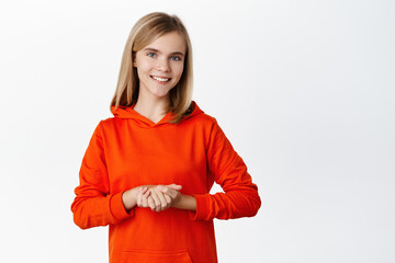 Portrait of cute smiling schoolgirl, beautiful blond girl standing in polite pose, holding hadns together, assisting or helping, standing over white background