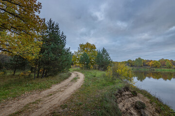 Fototapeta na wymiar Dirt road along the river bank. Cloudy autumn weather. Rural landscape with road, river and autumn forest.